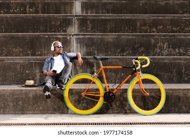 Happy hispanic man with blond hair sitting on stands and listening to music beside a fixie colorful bicycle. Eco-friendly urban transport concept.