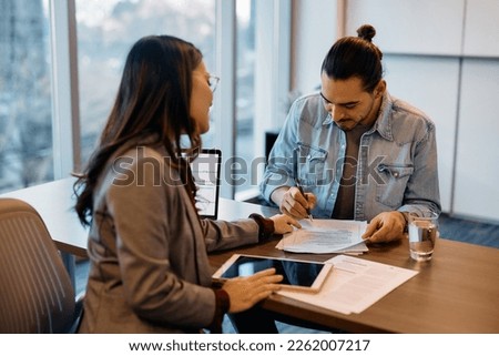 Happy Hispanic man signing paperwork while having meeting with insurance agent in the office.