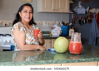 Happy Hispanic Mom Drinking Natural Watermelon Juice - Mature Woman Preparing Natural Watermelon Soda - Woman Drinking Delicious Fresh Juice In Glass At Table In Kitchen.