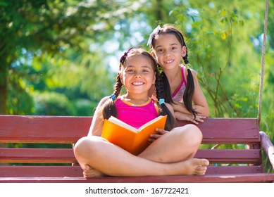 Happy Hispanic Sisters Reading Book In Summer Park