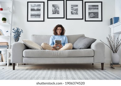 Happy Hispanic Teen Girl Holding Pad Computer Gadget Using Digital Tablet Technology Sitting On Couch At Home. Smiling Young Woman Using Apps, Shopping Online, Reading News, Browsing Internet On Sofa.