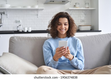 Happy Hispanic Teen Girl Looking At Smartphone Relaxing On Couch At Home, Enjoying Using Online Mobile Apps Technology, Playing Games On Cell Phone, Checking Messages Or Social Media Posts.