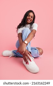 Happy Hispanic Teen Model In Trendy Clothes Embracing Knee And Touching White Sneaker While Sitting Against Pink Background And Smiling