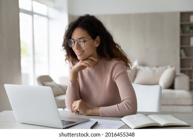 Happy Hispanic woman in glasses sit at desk work online on laptop make notes handwrite. Smiling Latino female student study on web on computer, take distant course or training on gadget at home. - Powered by Shutterstock