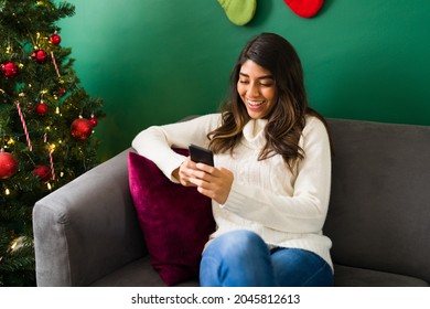 Happy Hispanic Woman Texting On Her Smartphone And Using Social Media While Waiting To Celebrate Christmas At Home 