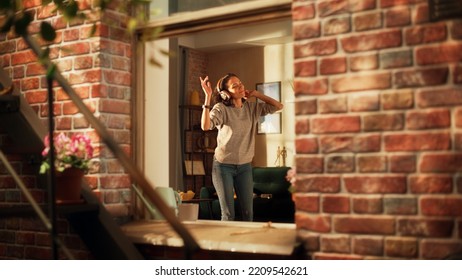 Happy Hispanic Woman Wearing Wireless Headphones And Dances In Her Cozy Apartment. Cheerful Lovely Girl Listening To Music At Home And Dancing In A Good Mood. Shot Throught Apartment Window.