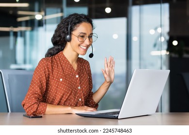 Happy hispanic woman working inside office, businesswoman with video call headset talking and advising customers remotely, tech support online store customer service, greeting with hand. - Powered by Shutterstock