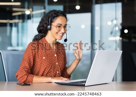 Happy hispanic woman working inside office, businesswoman with video call headset talking and advising customers remotely, tech support online store customer service, greeting with hand.