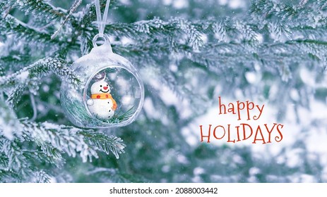 Happy Holidays greeting card. Christmas glass ball with cute snowman toy on snowy fir tree, natural background. Christmas and new year holidays. festive winter season - Powered by Shutterstock