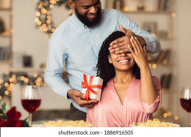 Happy Holidays. Portrait Of Smiling Black Man Covering His Woman Eyes And Giving Her Present Box, Making Surprise To Beautiful Lady. African American Couple Spending Time Together At Home Or Cafe