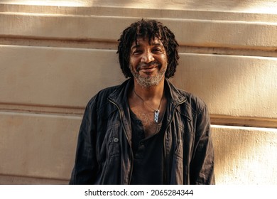 Happy homeless man smiling at the camera cheerfully while standing alone during the day. Self-confident homeless man standing against a brick wall outdoors in the city.