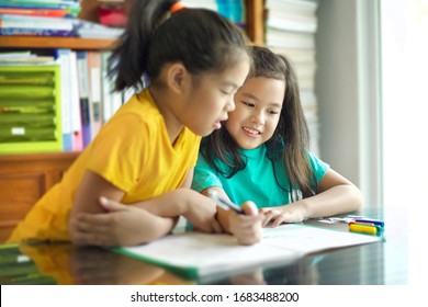 Happy homeschooled children doing homework.Beautiful Asian siblings laughing while homeschooling.Female child homeschooler writing with pen.Colorful crayons on table with shelves in background. Sister