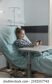 Happy Hospitalized Little Girl Watching Funny Cartoons On Smartphone While Resting Inside Healthcare Clinic Pediatric Ward Room. Sick Kid With Modern Mobile Phone Enjoying Funny Content On Internet.