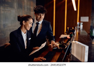 Happy hotel managers through reservation list on desktop PC at reception desk. 