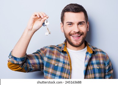 Happy House Owner. Smiling Young Man Holding Keys And Looking At Camera While Standing Against Grey Background