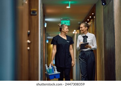 Happy housekeeping executive using touchpad while communicating with chambermaid in a hotel hallway. Copy space.