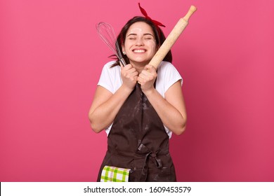 Happy housewife or baker chef wearing kitchen apron holding baking rolling pin, posing isolated over rose studio background, keeping eyes closed and stands with hands under chin, expresses happyness.