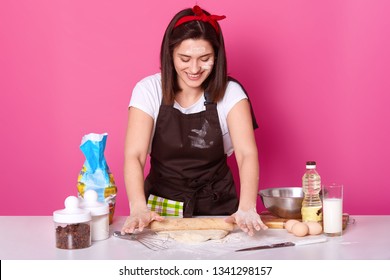 Happy Housewife Or Baker Wears Kitchen Apron Dirty With Flour, White T Shirt, Red Headband, Holds Baking Rolling Pin And Rolls Out Dough. Studio Picture Isolated Over Pink Background. Baking Concept.