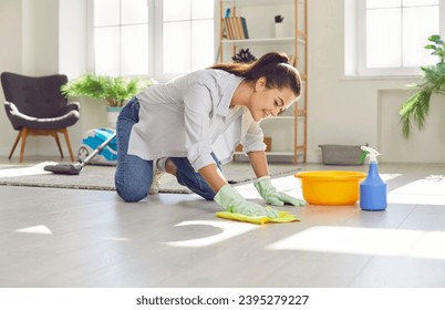 Happy housewife cleaning the floor at home. Young woman in rubber gloves washing and wiping light gray wooden or laminate flooring in the living room. Housework concept - Powered by Shutterstock