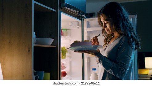 Happy hungry woman having a midnight snack, she is standing in front of the fridge and eating a delicious cake - Shutterstock ID 2199248611