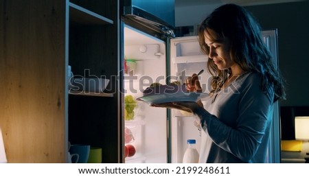 Happy hungry woman having a midnight snack, she is standing in front of the fridge and eating a delicious cake