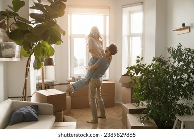 Happy husband lifts his young beautiful wife, laughing, swirling, enjoy move-in day to new, first house, big carton boxes nearby in unfurnished sunny living room. Bank loan for young family, new life - Powered by Shutterstock