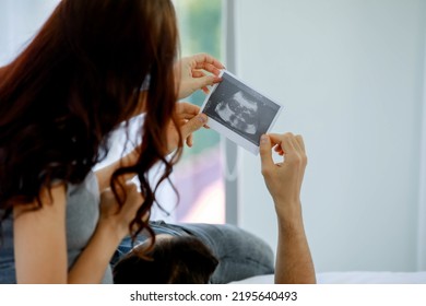 Happy Husband Lying Down On Hip Of Beloved Pregnant Wife And Looking At Paper Of To-do List And Discussion For Expecting Birth Of Unborn Baby. Young Couple Prepare Child Cloths On Bed At Sweet Home.