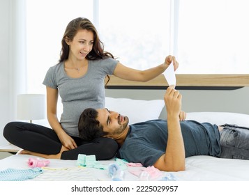 Happy Husband Lying Down On Hip Of Beloved Pregnant Wife And Looking At Paper Of To-do List And Discussion For Expecting Birth Of Unborn Baby. Young Couple Prepare Child Cloths On Bed At Sweet Home.