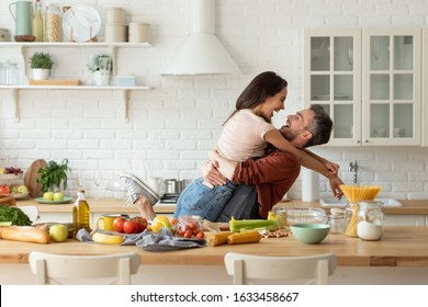 Happy Husband And Wife Having Fun During Cooking At Home Kitchen. Excited Man Hugging And Carrying Loving Woman, Cozy Bright Loft Interior With Table Full Of Fresh Healthy Groceries Food. Laughter Fun