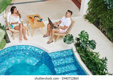 Happy Husband And Wife Sitting By Swimming Pool In Their Beach House And Working On Laptop And Digital Tablet