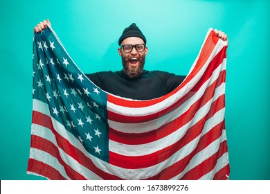 Happy Independence Day USA. Handsome American Hipster Man With United States Flag In Hands. Guy With Beard Wearing Casual Beanie Black Longsleeve Shirt