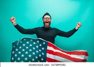 Happy Independence Day USA. Handsome American Hipster Man With United States Flag In Hands. Guy With Beard Wearing Casual Beanie Black Longsleeve Shirt