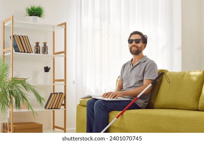 Happy independent disabled blind man in dark eyeglasses rests on sofa, holds Braille sign symbol system book, uses tactile sense, feels paper pages with fingers and reads textured dot-patterned script - Powered by Shutterstock