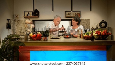 Happy Indian aged couple preparing vegetarian salad standing in modern kitchen at home. Senior wife in apron teaching husband to stir mix fresh vegan soup ingredients meal in pot with wooden spatula