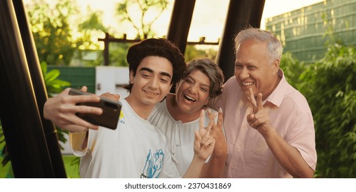 Happy Indian aged parents standing together teenager boy holding mobile phone taking funny faces selfie making fun outdoor house. Diverse family looking camera enjoying summer vacation at outside park - Powered by Shutterstock