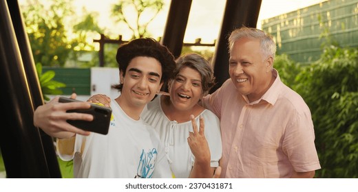 Happy Indian aged parents standing together teenager boy holding mobile phone taking funny faces selfie making fun outdoor house. Diverse family looking camera enjoying summer vacation at outside park - Powered by Shutterstock