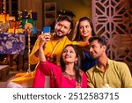 Happy Indian Asian couples and friends in traditional wear celebrating Diwali at home with drinks, sweets, gifts, flowers, taking selfies, and enjoying together.