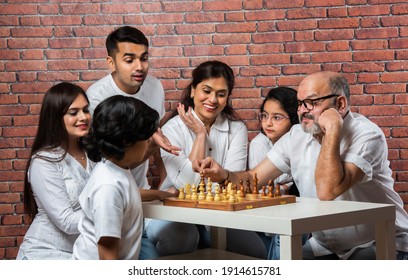 Happy Indian Asian Multigenerational Family Of Six Playing Chess Or Shatranj Which Is A Popular Board Game, Wearing White Cloths Against Red Brick Wall