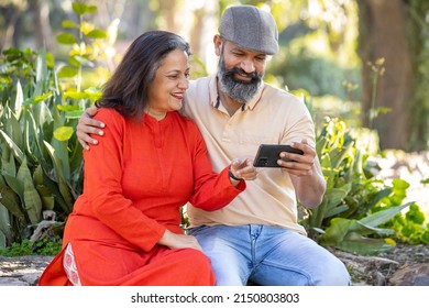 Happy Indian Asian Senior Couple Using Smart Phone At Park, Old Man And Woman Watching Video Social Media On Mobile Phone While Sitting On Bench In The Garden.