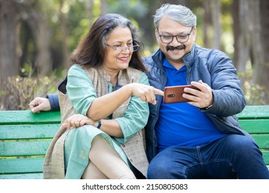 Happy Indian Asian Senior Couple Using Smart Phone At Park, Old Man And Woman Watching Video Social Media On Mobile Phone While Sitting On Bench In The Garden.