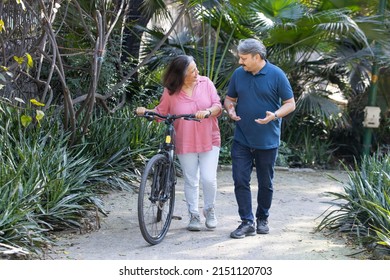 Happy Indian Asian Senior Couple Walking Their Bike Talking In Park.mature Couple In Summer Park.Retired Man And Old Woman With Bicycles Outside Nature.