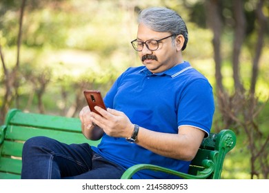 Happy Indian Or Asian Senior Man Using Chatting On Smart Phone While Sitting On The Bench At Park , Old Grey Hair Male Wearing Glasses Relaxing Using Mobile Phone Outdoor, Social Media  Technology.