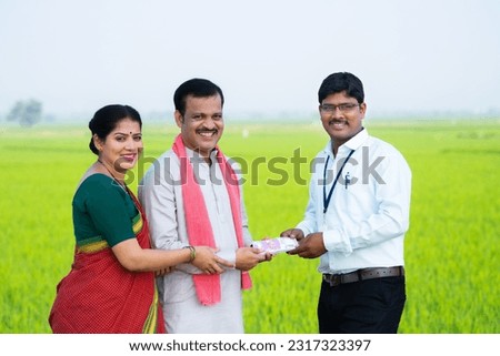 Happy indian banker giving money to farmer village couple by looking at camera at farmland - concept of agribusiness, financial helping and banking support