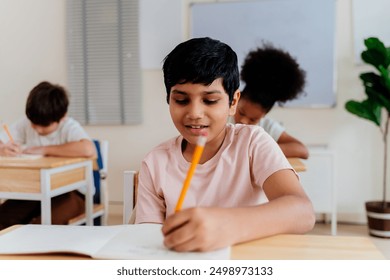 Happy Indian boy in a classroom setting, smiling brightly while writing. Young student with afro hair, sitting at a desk with a pencil and notebook - Powered by Shutterstock