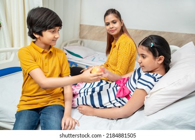 Happy indian brother give apple to his sister at hospital bed. little sick girl lying in bed with her mother sitting by her side at clinic. children health insurance - Shutterstock ID 2184881505