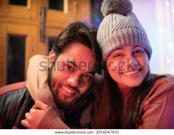 Happy Indian brother and sister sitting together\
at home in winters and embracing each other with a toothy smile and\
looking at the camera. They are wearing warm clothes, sweaters,\
jacket andcap.