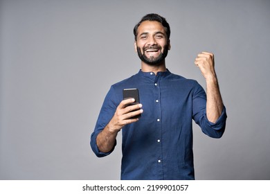 Happy indian business man investor winner using cell mobile phone isolated on gray background. Excited ethnic guy holding smartphone feels surprised about betting win on cellphone, celebrating success