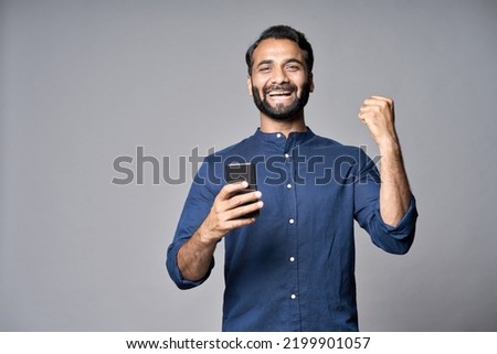 Happy indian business man investor winner using cell mobile phone isolated on gray background. Excited ethnic guy holding smartphone feels surprised about betting win on cellphone, celebrating success