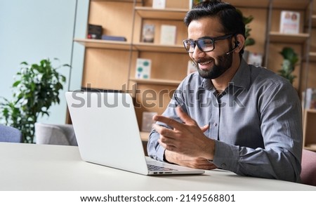 Happy indian business man remote teacher, customer support manager wearing headset talking at virtual meeting consulting client on video call giving distance learning class at home office call center.