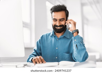 Happy indian businessman in blue shirt talking on smartphone, checking schedule in planner on desk in bright office setting, free space - Powered by Shutterstock
